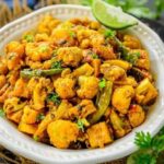 Dish of ALO GOBI with potatoes, cauliflower, ginger, garlic, and cumin garnished with lime and cilantro.