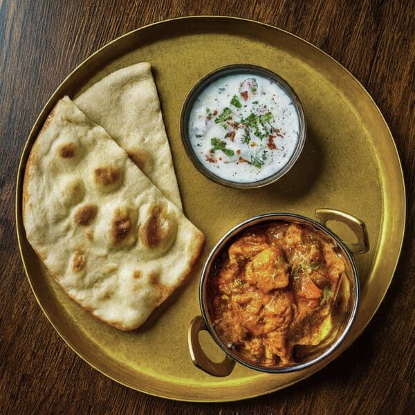 Chicken Korma with coconut, cream, and cashew sauce served with naan bread and raita on a wooden table.