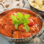 A bowl of Tikka Masala with a garnish of fresh herbs, served alongside rice and bread.