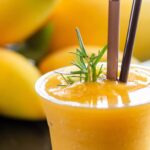 Refreshing mango lassi in a clear glass with straws, garnished with rosemary, surrounded by fresh mangoes.